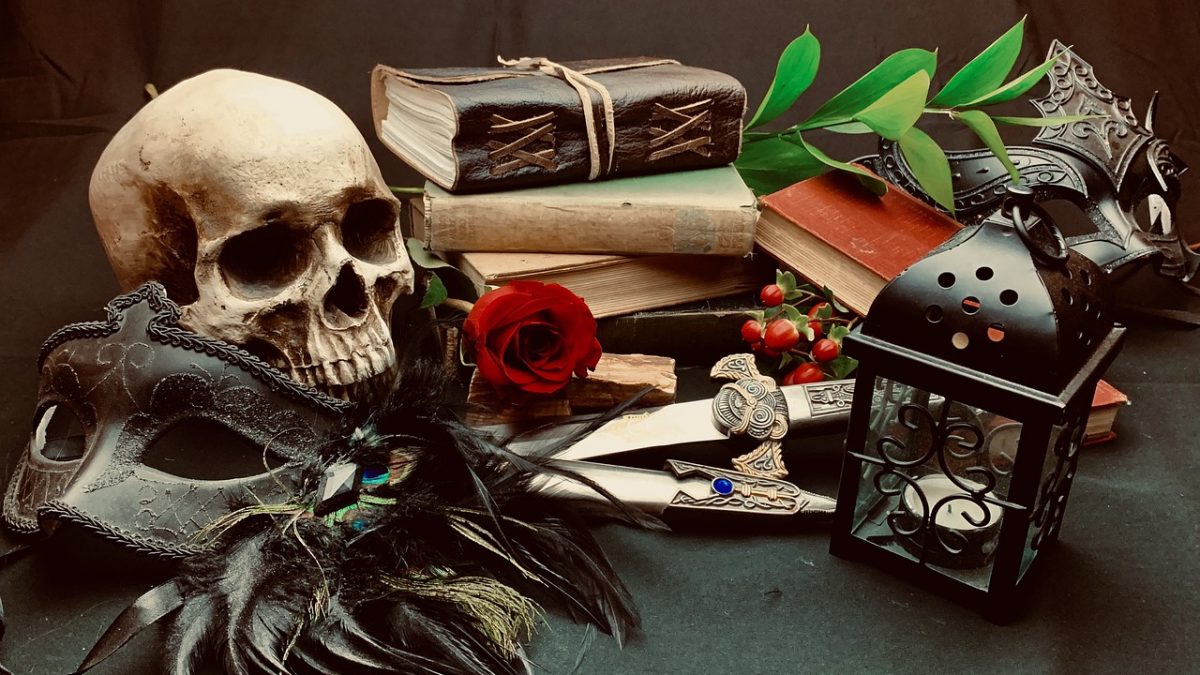 A plastic human skull with a feathered opera mask to the left of a stack of books, cherries, swords, and a lantern.