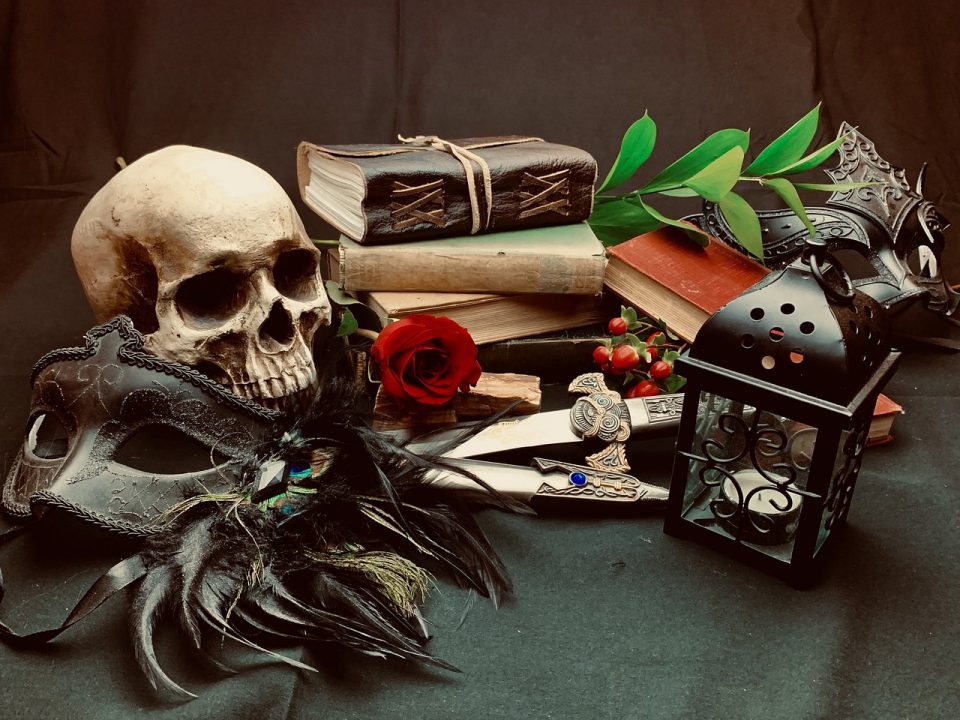 A plastic human skull with a feathered opera mask to the left of a stack of books, cherries, swords, and a lantern.